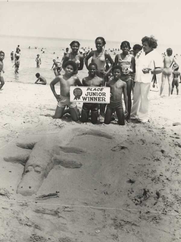 history photo of six Black children in swimsuits at South Shore Beach holding up a 'third place winner' sign next to their sand sculpture of a crocodile. Behind them are many swimmers of all ages standing on the beach and in the water.